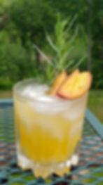 An orange cocktail with 3 slices of peach and a sprig of tarragon in it, sitting on a table outside.