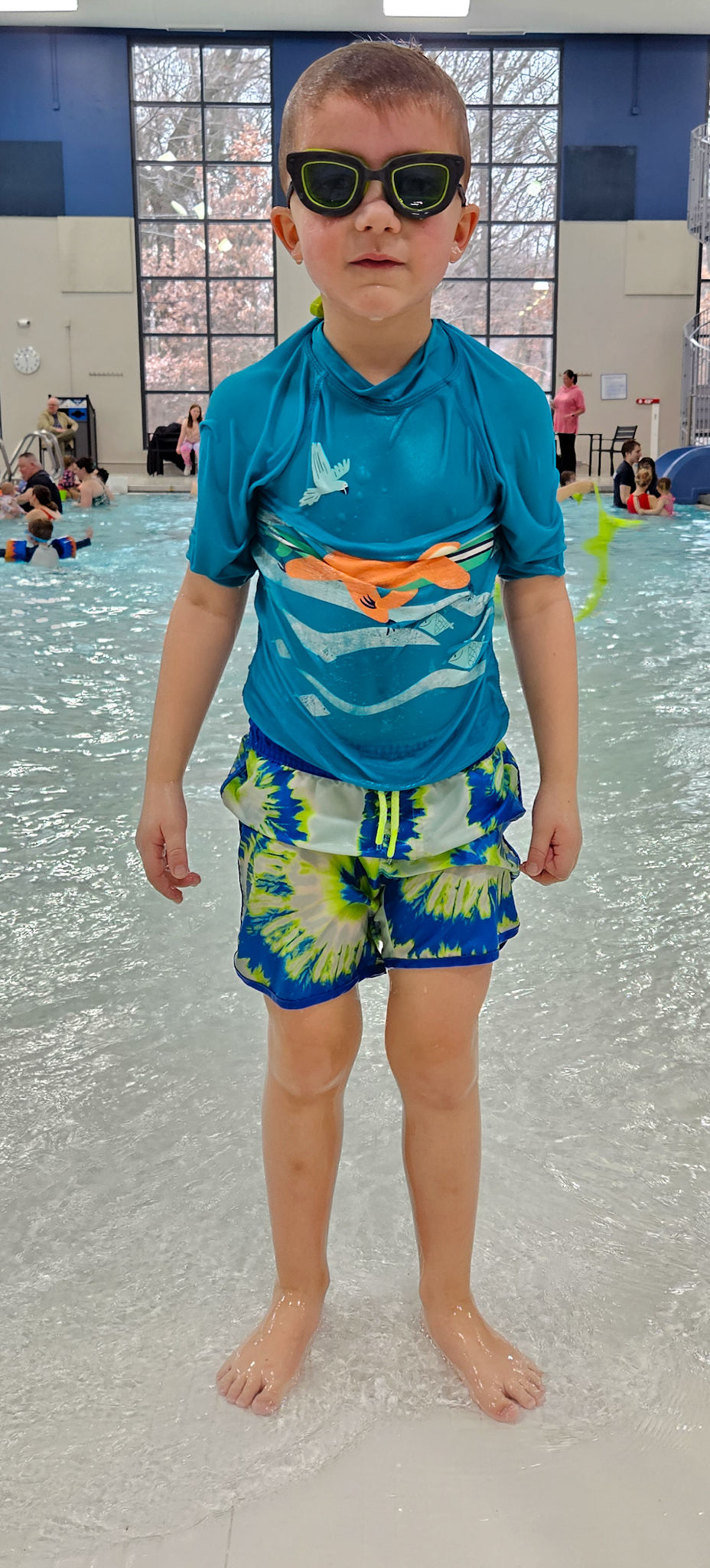 A boy in blue and green tie dye swim trunks, a a blue swim shirt, and black goggles standing by a public swimming pool with other swimmers in the background.