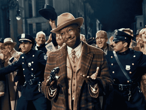 Lamorne Morris as Robbie Robertson smiles while holding a camera in Spider-Noir.