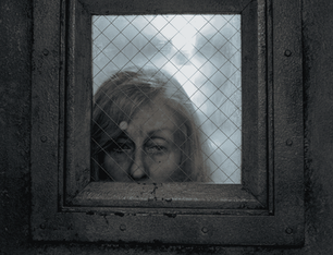 Close-up of actor Judith Light with grey hair and a weary expression, looking through a small reinforced window in a dark metal door.