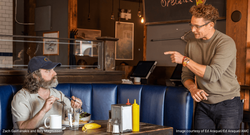 In a scene from the 2026 production The Audacity, actors Zach Galifianakis and Billy Magnussen are shown in a dimly lit diner. Galifianakis, with a beard and wearing a blue baseball cap and white t-shirt, sits in a blue booth holding a fork. Magnussen stands over him, wearing glasses and a green sweater, pointing a finger while appearing to be in the middle of a conversation.