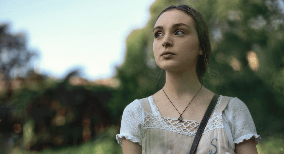 Chloe Jean Lourdes as Sarah Reid stands outdoors wearing a cream shirt marked with the letter S in the Apple TV thriller Last Seen.