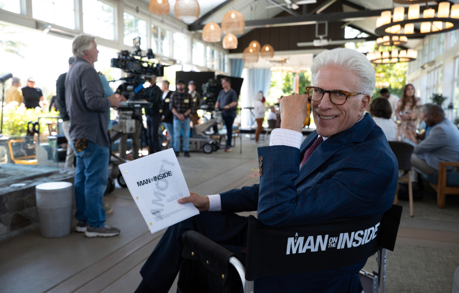 Ted Danson smiles in a blue suit and glasses, holding a script while seated near a camera crew on the A Man on the Inside set.