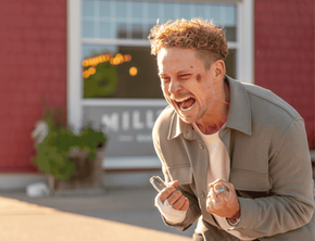 Actor Billy Magnussen portrays an intensely emotional scene in the series The Audacity. He is captured mid-shout with a strained expression, holding a pair of eyeglasses in his hand. He has a small visible bruise or mark on his cheek and is wearing a tan, button-down shirt.
