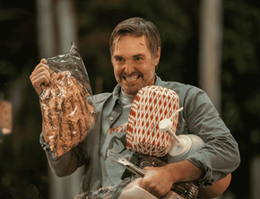 Will Forte as Jack smiles outdoors, holding a clear bag of buns, a large plastic water jug, and other various supplies.