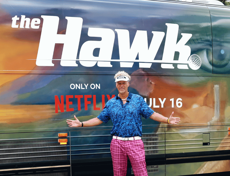 Will Ferrell stands in front of a bus as Lonnie The Hawk Hawkins wearing a blue polo shirt and pink plaid pants.