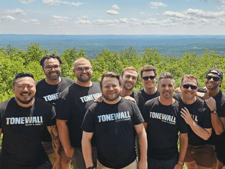 A group of 11 diverse men in matching TONEWALL t-shirts stand in a line on a grassy hill overlooking a vast, hilly landscape. 