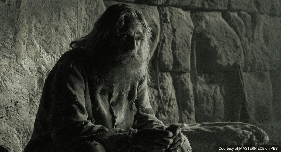 A close-up of actor Jeremy Irons as Abbé Faria in a dimly lit stone prison cell. He has long, unkempt grey hair and a thick beard, wearing a tattered, dirt-stained tunic while looking off-camera with a weary, soulful expression.