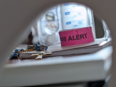 A vignette through the side of a hospital bed showing a rosary and a limb alert wrist band.