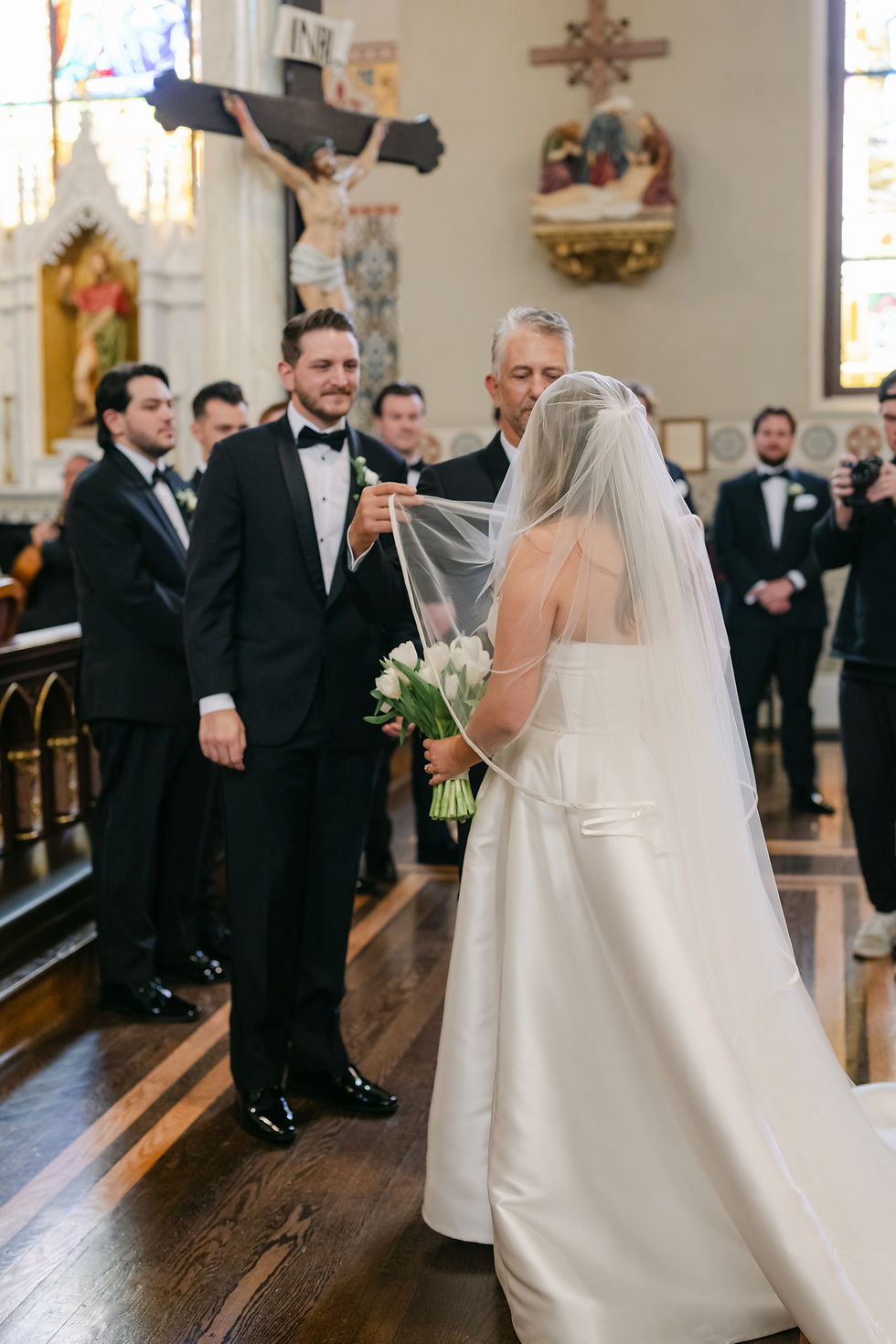 Father of the bride lifting the bride’s veil as her groom smiles waiting to exchange vows.