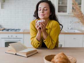 Woman in yellow shirt enjoys a cup of coffee in a bright kitchen. Open book and croissants are on the table. She looks relaxed and content.