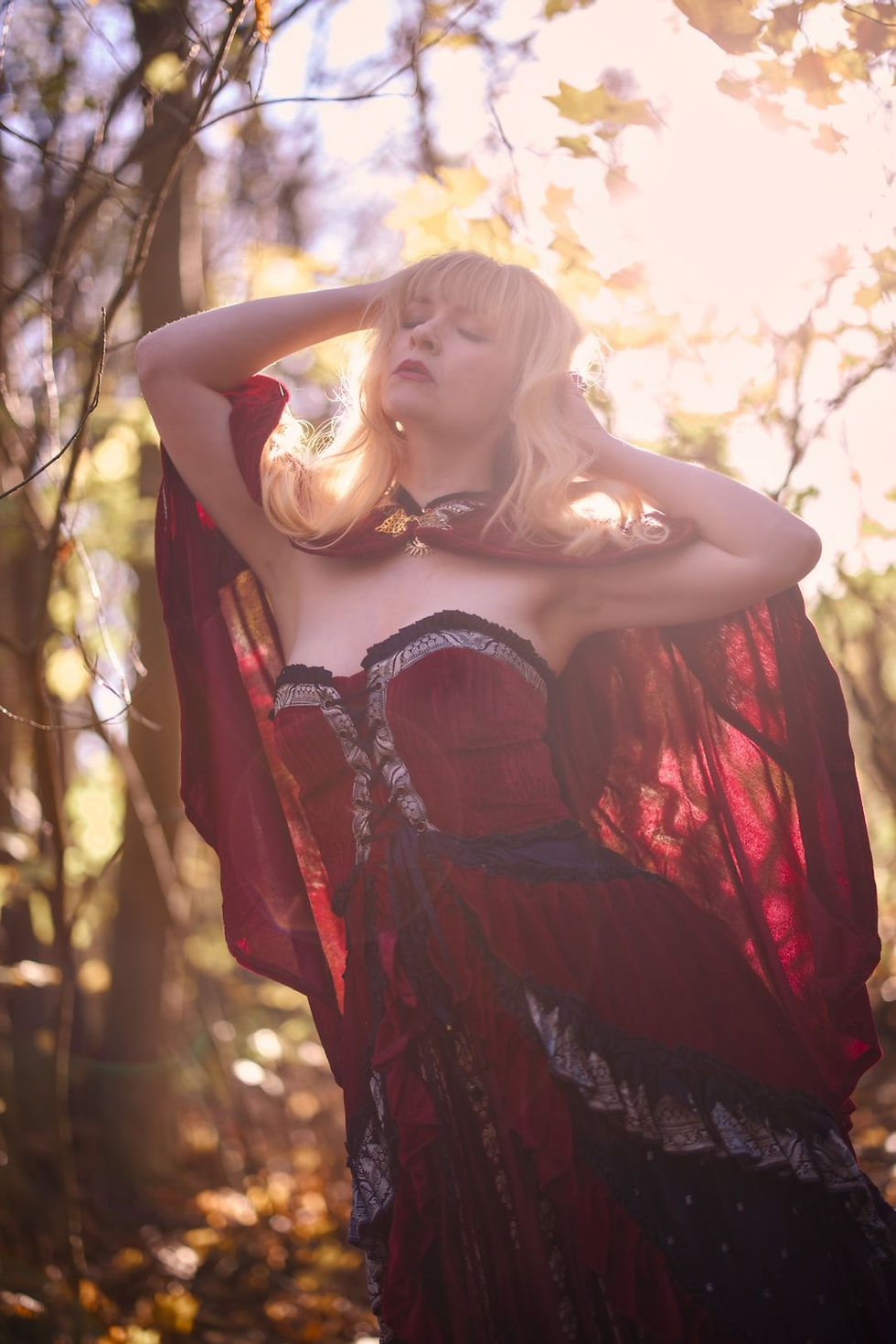 A blonde woman in a corseted red gown and red cape stretches in golden forest light, with her hands in her hair