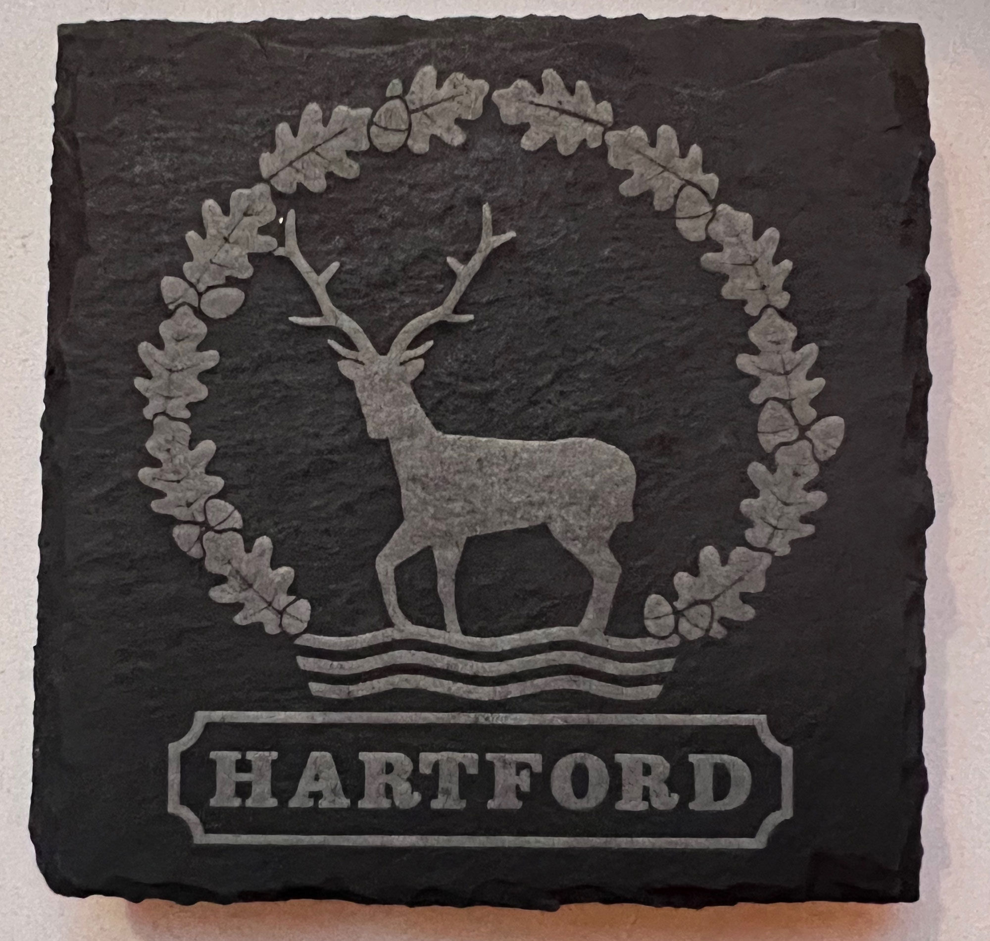 A square drinks coaster, made of dark grey slate, engraved with the Hartford village symbol of a Hart crossing a river