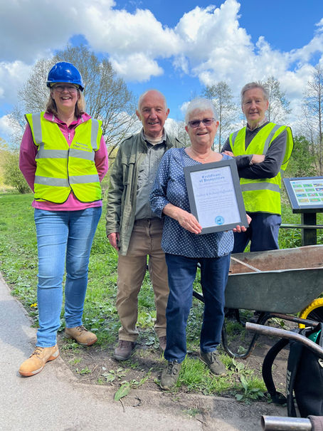 A framed Civic Award certificate is presented to two of Marshall's Arm Nature Reserve's longest standing supporters. One of the Reserve's meadow areas can be seen in the background.