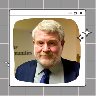 Dan is a white man with white hair and facial hair in a blue suit and tie. He is looking at the camera and smiling.