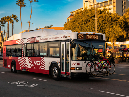 Red and white MTS bus labeled "923 Downtown" with two bikes on front, drives on a sunlit street lined with palm trees and cafes.