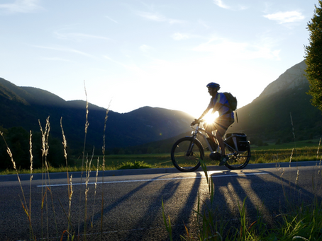 Cyclist rides on a road at sunrise, casting long shadows. Mountains and trees form the tranquil background, with a clear blue sky.