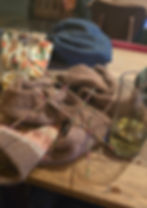 Knitting project on a wooden table with colorful needles, beer glasses, a floral pouch, and a blue cap. Cozy and relaxed setting.