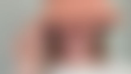 Woman smiling, wearing a peach hat and white shirt, tilts hat playfully. She has long hair and a gold bracelet. Neutral background.