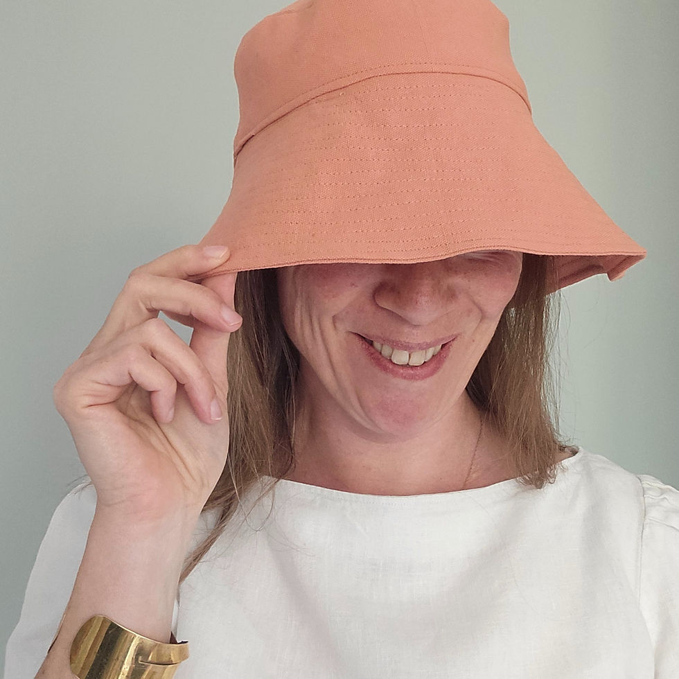 Woman smiling, wearing a peach hat and white shirt, tilts hat playfully. She has long hair and a gold bracelet. Neutral background.