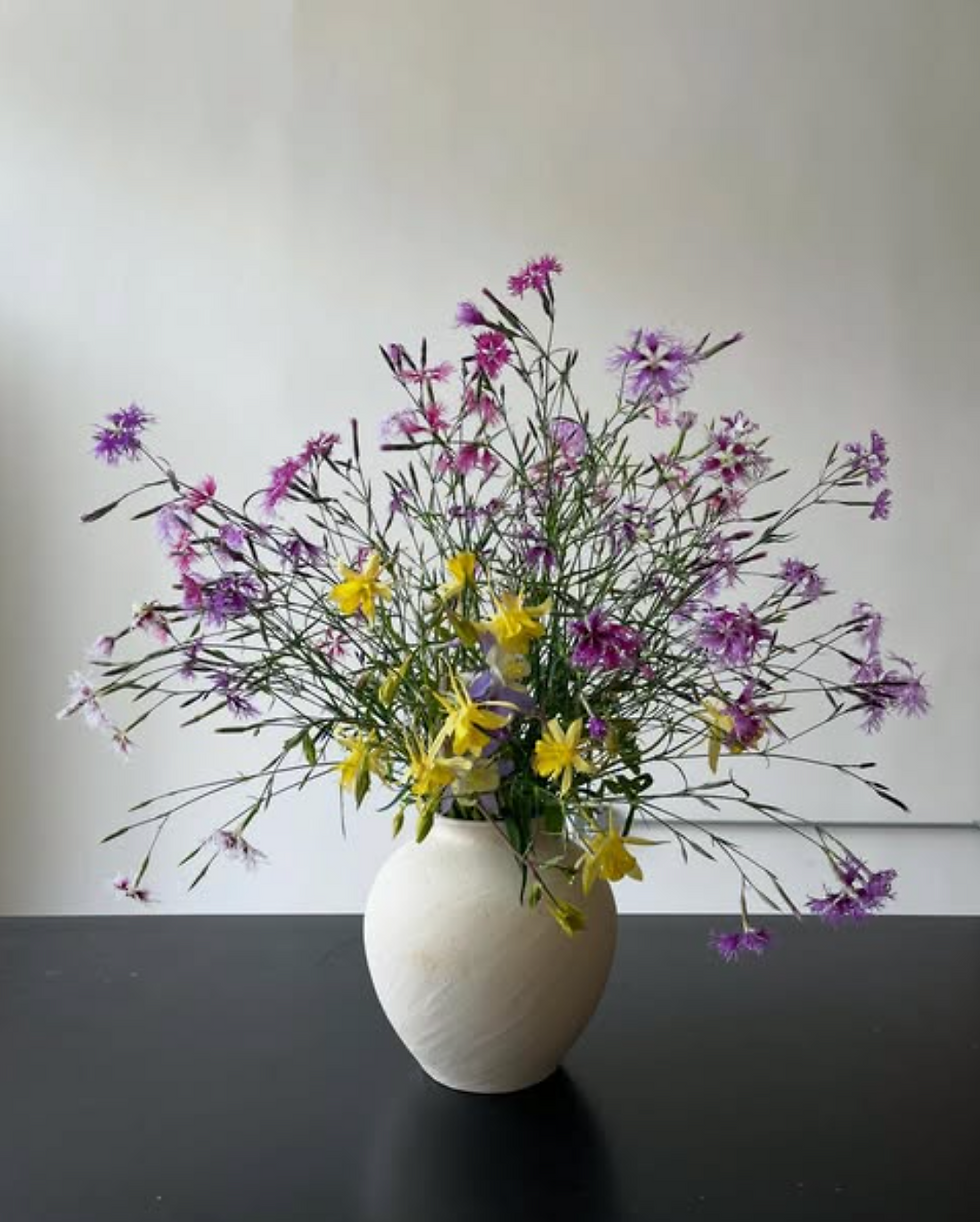 A white vase holds vibrant purple and yellow wildflowers, set against a plain, light backdrop on a dark surface, creating a serene mood.