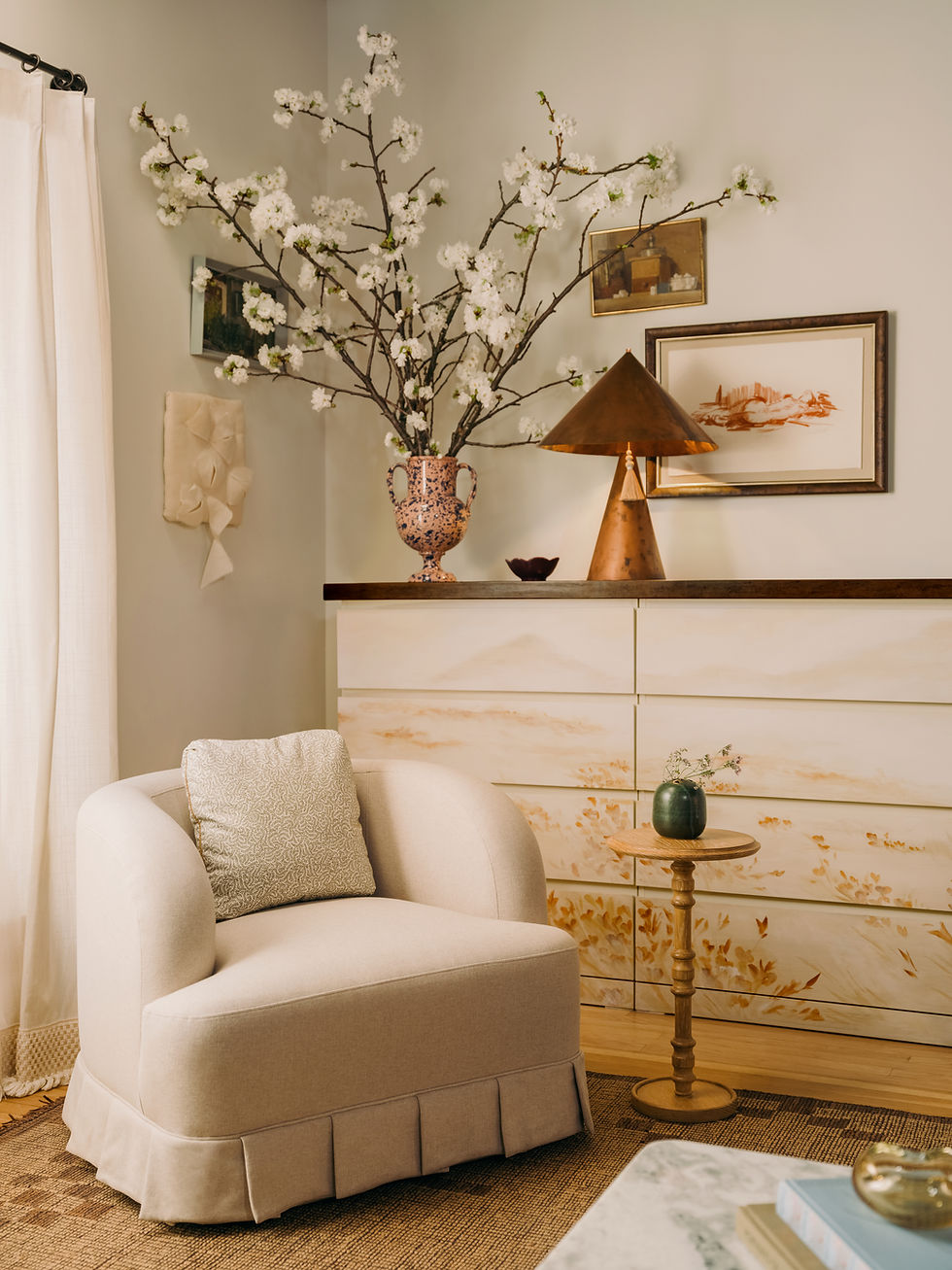 Cozy room with skirted and pleated ivory chair with oak side table in front of a mural painted dresser in the background, styled with vase with branching florals and copper table lamp, paired with vintage works of art.