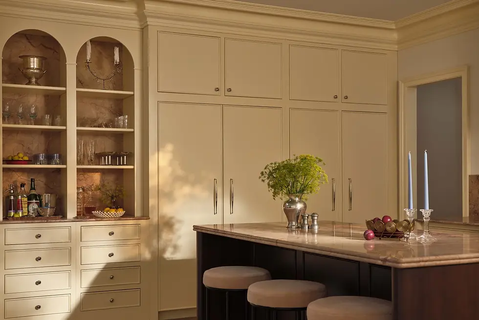 Elegant kitchen with cream cabinets, marble counters, three stools. Silver vase with greenery, blue candles, and fruit bowl on island. Warm light.