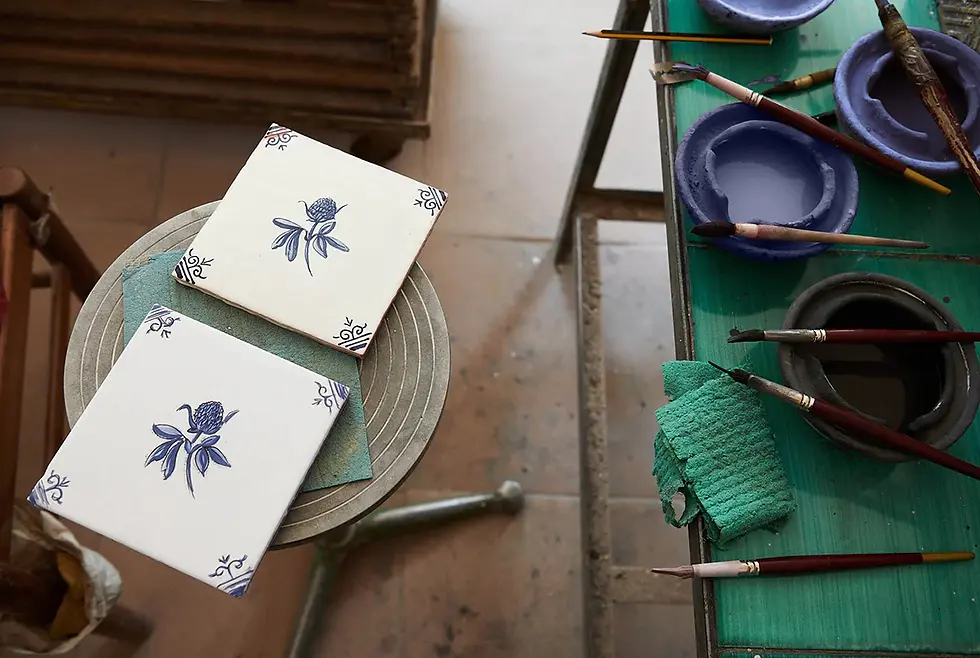Two white tiles with blue floral designs sit on a stool beside paintbrushes and bowls of paint on a green table. Artistic workshop setting.