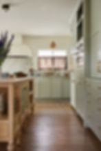 Cozy kitchen with celedon green cabinets, marble backsplash, and wooden island. Purple flowers on the counter. Warm, inviting atmosphere.