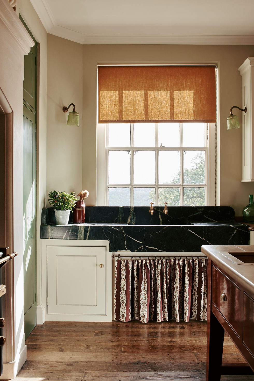 Vintage kitchen with a green marble sink, brass faucet, and wooden counter. Sunlit window with orange blind, potted plant, and rustic vibe.