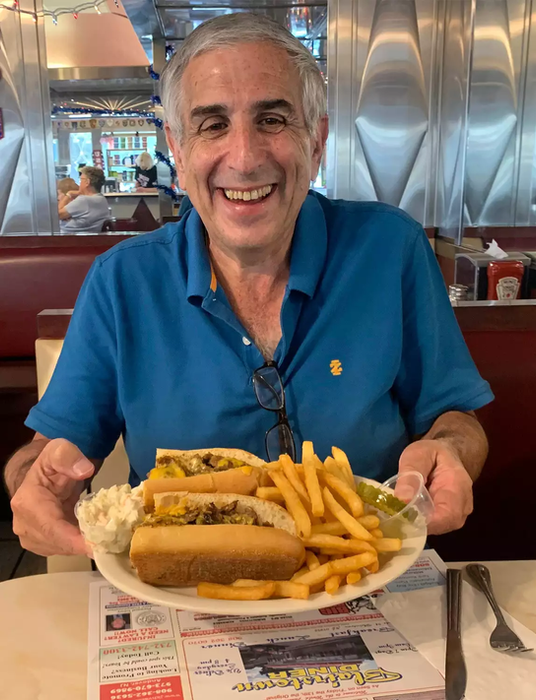 Smiling man holds two delicious-looking sandwiches, fries, and coleslaw at a diner.