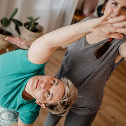 Yoga-Trainer unterstützt Frau bei einer Yoga-Übung. Beide lächeln, grünes Shirt, graues Shirt.
