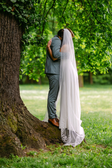 Brautpaar umarmt sich unter einem alten Baum im Park, Braut mit langem Schleier und Spitzenkleid
