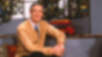 Mister Rogers sitting in a chair, wearing a tie and a yellow cardigan