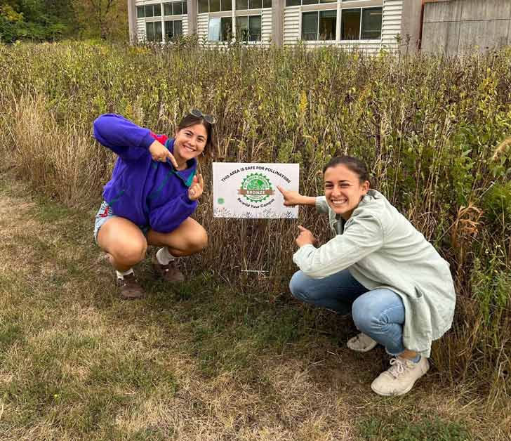 Mackenzie Feldman and Bridget Gustafson kneeling down next to a Green Grounds certification sign