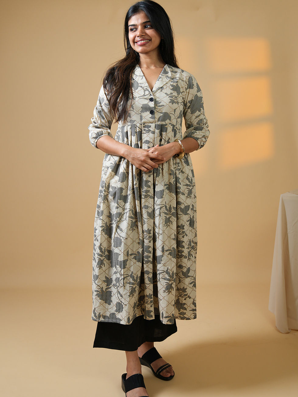 Woman smiling, wearing a cream patterned kurta, black pants, and sandals.