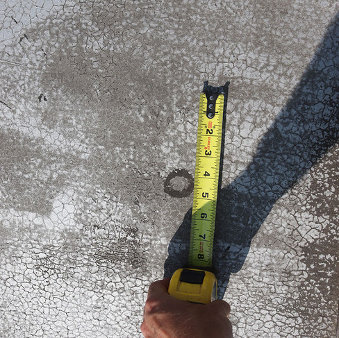 Photo of a person's hand holding a measuring tape next to a dark circular area on a rooftop.