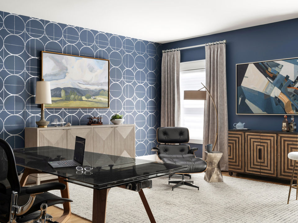 A contemporary home office in the Whitewater area, featuring a glass-topped desk with a laptop and eyeglasses on it. The room is decorated with a blue accent wall displaying a geometric circle pattern, complemented by an abstract landscape painting. A black leather lounge chair with an ottoman sits beside a modern floor lamp and a small sculpted side table. A light wood credenza and gray curtains add to the sophisticated and stylish decor.