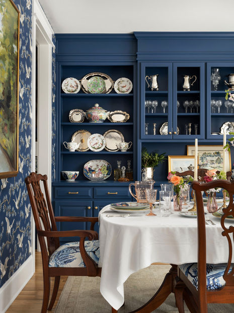 The dining table, covered with a white tablecloth, is set with elegant china, crystal glassware, and a floral centerpiece. Classic wooden chairs with blue and white patterned cushions surround the table. The room’s walls are adorned with blue and gold patterned wallpaper, complementing the sophisticated and timeless decor.
