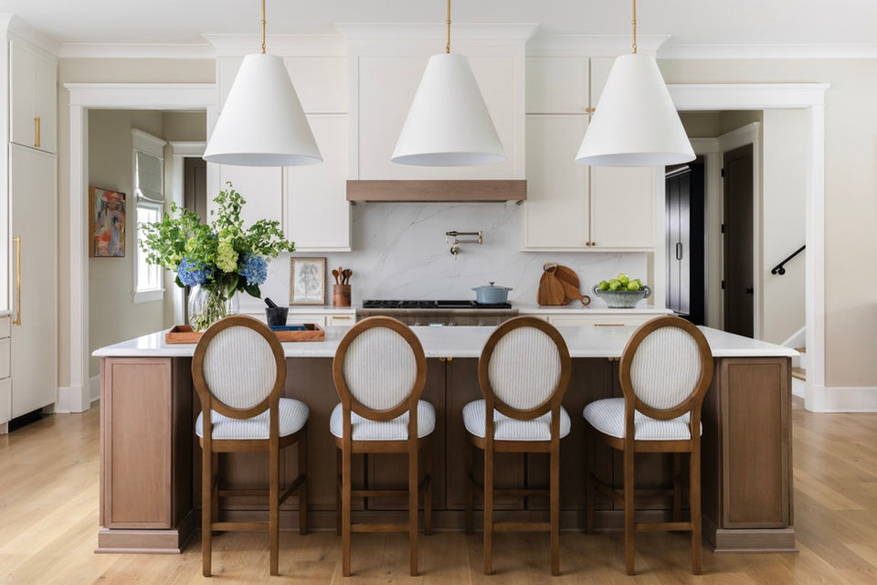Custom kitchen design in Senoia, GA showcasing a marble waterfall island, upholstered counter stools with wood frames, brass-accented white cabinetry, trio of white pendant lights, and a marble backsplash with warm wood detailing.