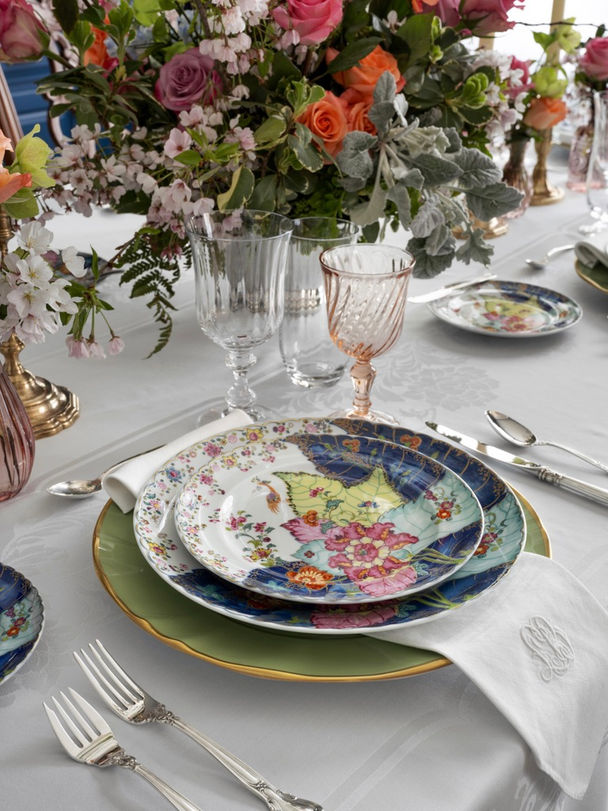 A beautifully set dining table located in the Whitewater area, designed by a Peachtree City-based interior designer. The table features an elegant arrangement of floral-patterned china plates stacked atop each other, each with intricate and colorful designs. The place setting includes a large green charger plate with a gold rim, a middle plate with blue and pink floral patterns, and a top plate with delicate multicolored flowers. Silver cutlery is neatly arranged alongside a monogrammed white napkin. Crystal glassware, including a clear wine glass and a blush-tinted goblet, add a touch of sophistication. The table is adorned with a lush centerpiece of fresh flowers in vibrant hues of pink, orange, and lavender, complemented by greenery, enhancing the table's luxurious and inviting ambiance.