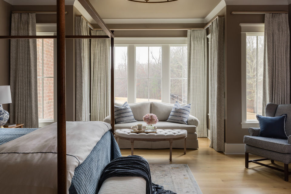 Light-filled primary bedroom sitting area in Senoia, GA featuring a curved loveseat, striped blue accent pillows, tufted ottoman coffee table, and floor-to-ceiling drapery in soft neutral tones.