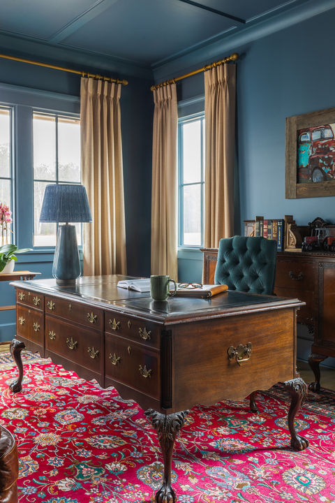 Moody blue home office in Senoia, GA featuring a traditional wood executive desk, red Persian-style rug, tailored drapery, tufted accent chair, and layered antique-inspired furnishings.
