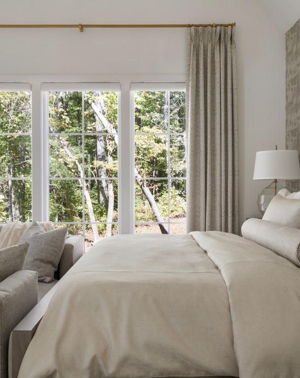 A bright bedroom with a beige bed, gray sofa, large windows with cream curtains, and a view of a green forest. Interior Design project in the Serenbe community in Chattahoochee Hills, Georgia.