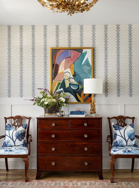 A traditionally styled interior in the Whitewater area, showcasing a dark wood chest of drawers flanked by two classic chairs with blue and white botanical-patterned cushions. Above the chest hangs a vibrant abstract painting in a gold frame. The room features wallpaper with a vertical leaf pattern and wainscoting on the lower half of the wall. Decorative items on the chest include a table lamp with a gold leaf base and a vase of fresh flowers, all under an ornate, gold-toned chandelier.