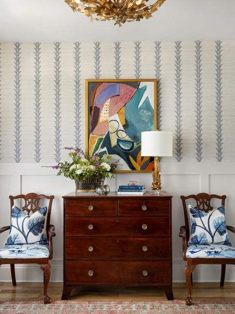 A traditionally styled interior in the Whitewater area, showcasing a dark wood chest of drawers flanked by two classic chairs with blue and white botanical-patterned cushions. Above the chest hangs a vibrant abstract painting in a gold frame. The room features wallpaper with a vertical leaf pattern and wainscoting on the lower half of the wall. Decorative items on the chest include a table lamp with a gold leaf base and a vase of fresh flowers, all under an ornate, gold-toned chandelier.
