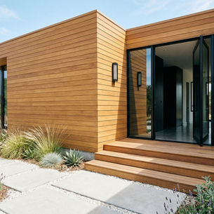 Exterior of a modern minimalist home clad in warm horizontal cedar wood siding with a black-framed glass pivot door, wood deck steps, a black wall-mounted light, and a drought-tolerant front landscape of concrete stepping stones, white gravel, and ornamental grasses under a clear blue sky.