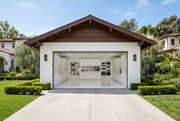 Front exterior view of a luxury detached two-car garage in a Los Angeles Mediterranean estate with white smooth stucco, a dark walnut wood gabled roof with exposed rafter tails, and the door fully open to reveal a pristine white interior with floor-to-ceiling custom white cabinetry with gold hardware, a neatly organized tool pegboard wall, and glossy white epoxy floors, flanked by manicured boxwood hedges, olive trees, and lush green lawn under a clear blue sky.