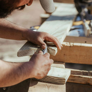 Close-up of a bearded carpenter's hands using a chisel to cut a precise wood joint notch into a pine lumber board on an outdoor workbench, with wood shavings visible and power tools blurred in the background in warm natural sunlight.