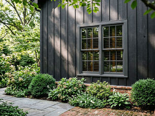 Close-up exterior of a home with dark charcoal black vertical board and batten siding, a gray-framed double-hung window with plantation shutters, lush green shrubs along a red brick border, and a bluestone pathway in the foreground.